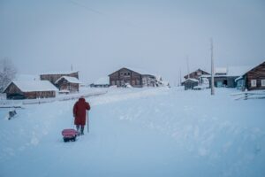 A wide shot of a village and a person in a red coat walking through the thick snow on a cold day